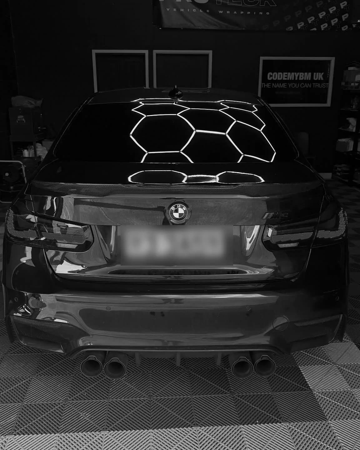 The CODEMYBM workshop F80 M3 from behind, under hexagon ceiling lights, with the CODEMYBM UK sign on the wall