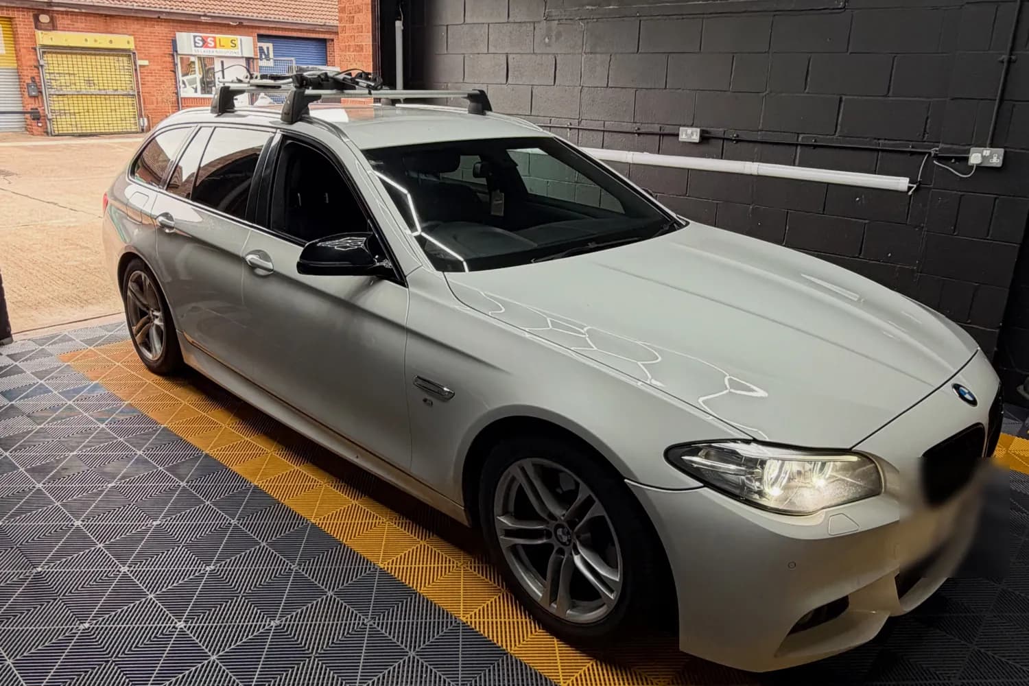 A white BMW F11 5 Series Touring in the CODEMYBM workshop bay, on yellow and grey floor tiles