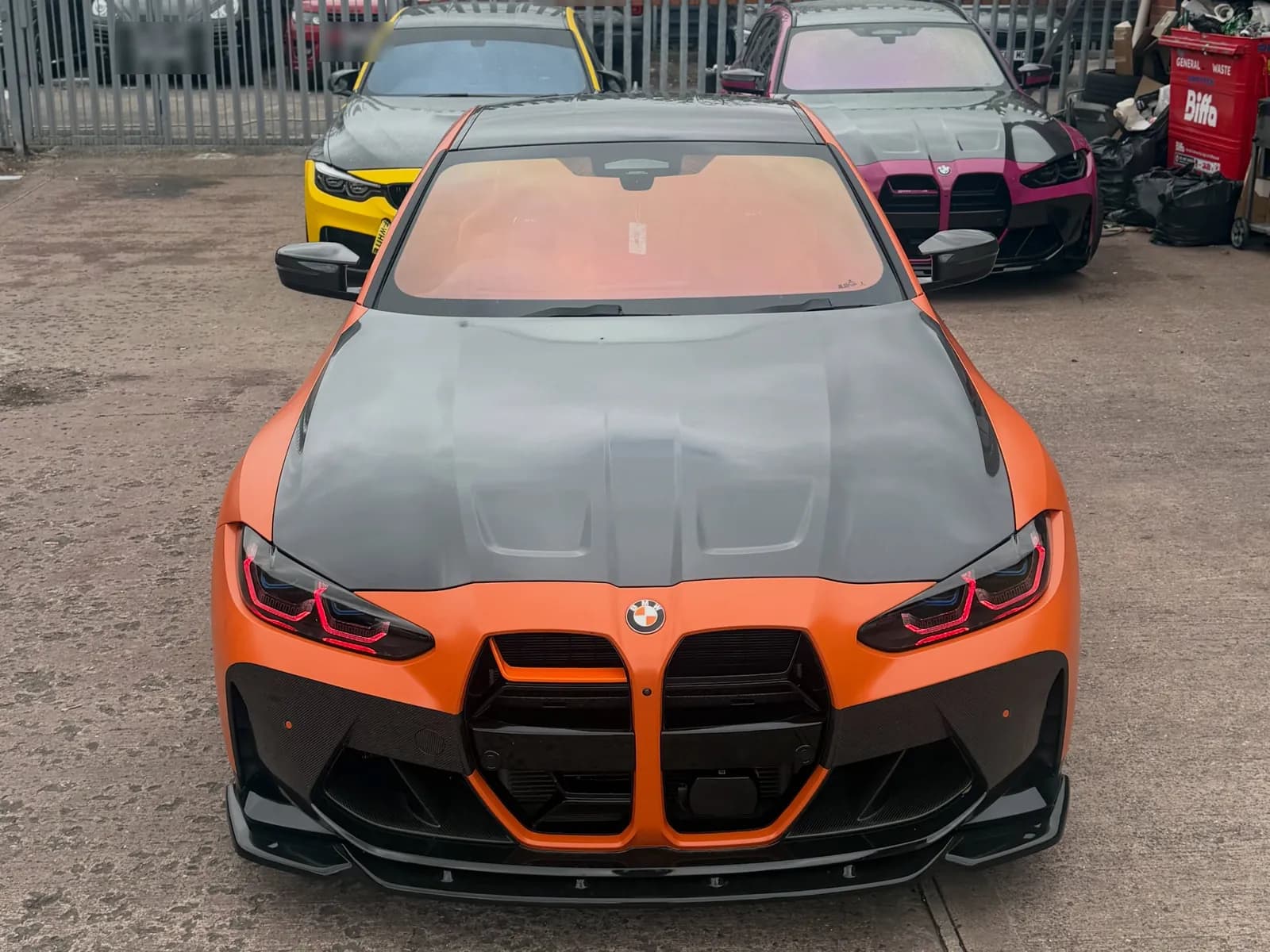 An orange BMW G82 M4 with a carbon bonnet outside the CODEMYBM workshop, with a yellow M4 and a pink M3 behind