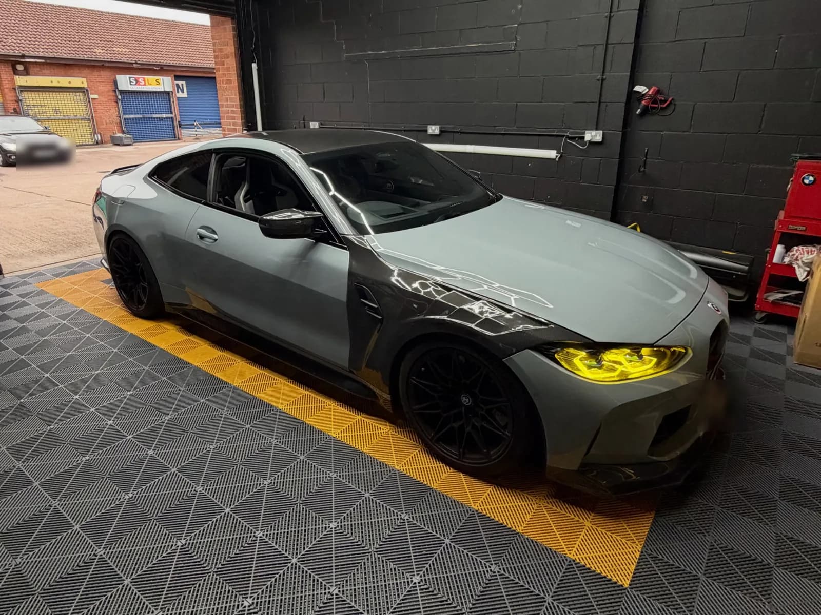 A grey BMW G82 M4 in the CODEMYBM workshop bay, on yellow and grey floor tiles